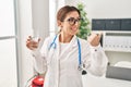Young brunette doctor woman holding glass of water pointing thumb up to the side smiling happy with open mouth Royalty Free Stock Photo