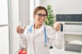 Young brunette doctor woman holding glass of water smiling happy pointing with hand and finger to the side Royalty Free Stock Photo