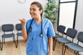 Young brunette doctor woman at waiting room smiling with happy face looking and pointing to the side with thumb up Royalty Free Stock Photo