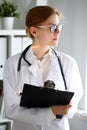 Young brunette female doctor standing with clipboard near window in hospital. Physician ready to examine patient Royalty Free Stock Photo