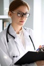 Young brunette female doctor standing with clipboard near window in hospital. Physician ready to examine patient Royalty Free Stock Photo