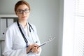 Young brunette female doctor standing with clipboard near window in hospital. Physician ready to examine patient Royalty Free Stock Photo