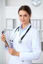 Young brunette female doctor standing with clipboard and smiling at hospital. Physician ready to examine patient Royalty Free Stock Photo