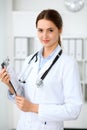 Young brunette female doctor standing with clipboard and smiling at hospital. Physician ready to examine patient Royalty Free Stock Photo