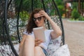 Young brunette girl in denim shorts white top black glasses. Resting in a chair on the outdoor in the hands of a tablet. Royalty Free Stock Photo