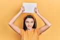 Young brunette girl holding blank empty banner over head smiling looking to the side and staring away thinking