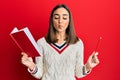 Young brunette girl holding book and pencil making fish face with mouth and squinting eyes, crazy and comical