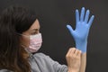 A young brunette girl puts on her left hand a blue medical disposable glove. A woman is wearing a protective mask. Dark background