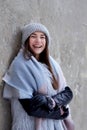 Young brunette girl, wearing grey fur coat and knit hat, leaning on grey concrete wall, smiling, laughing. Winter female portrait Royalty Free Stock Photo