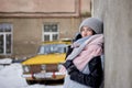 Young brunette girl, wearing grey fur coat and knit hat, leaning on grey concrete wall, with old yellow car on background.Winter Royalty Free Stock Photo