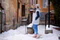 Young brunette girl, wearing grey fur coat and knit hat, standing in front of old yellow historical building. Winter female