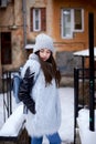 Young brunette girl, wearing grey fur coat and knit hat, standing in front of old yellow historical building. Winter female