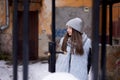 Young brunette girl, wearing grey fur coat and knit hat, standing in front of old yellow historical building. Winter female