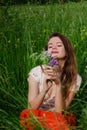 Young brunette girl in white top and red skirt sitting in green field grass in countryside smelling small bouquet of field flowers Royalty Free Stock Photo