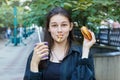 Young brunette holds a hamburger and a glass with a drink in her hands. French fries are clamped in her teeth