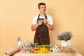 Young brunette man florist wearing brown apron standing in floral shop smelling rose anjoying aromat making order for customer Royalty Free Stock Photo