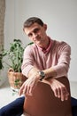 Young brunette man, wearing casual light pink jumper, sitting on brown stool in light room, thinking. Serious emotions.Three-