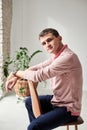 Young brunette man, wearing casual light pink jumper, sitting on brown stool in light room, thinking. Serious emotions.Three-