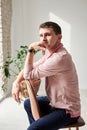 Young brunette man, wearing casual light pink jumper, sitting on brown stool in light room, thinking. Serious emotions.Three-