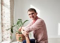 Young brunette man, wearing casual light pink jumper, sitting on chair by brick wall in light room with green plant, smiling,