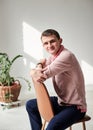 Young brunette man, wearing casual light pink jumper, sitting on chair by brick wall in light room with green plant, smiling,