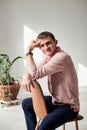 Young brunette man, wearing casual light pink jumper, sitting on chair by brick wall in light room with green plant, smiling,