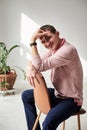 Young brunette man, wearing casual light pink jumper, sitting on chair by brick wall in light room with green plant, smiling,