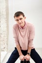 Young brunette man, wearing casual light pink jumper, sitting on chair by brick wall in light room. Serious emotions.Three-quarter
