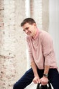 Young brunette man, wearing casual light pink jumper, sitting on chair by brick wall in light room, smiling, laughing. Happy