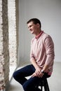 Young brunette man, wearing casual light pink jumper, sitting on chair by brick wall in light room, smiling, laughing. Happy