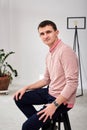 Young brunette man, wearing casual light pink jumper, sitting on chair by brick wall in light room. Serious emotions.Three-quarter