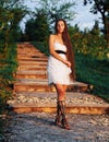 Young brunette model at the white dress staying on the stairs with green trees  in full growth Royalty Free Stock Photo