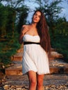 Young brunette model at the white dress staying on the stairs with green trees  posing and looking at the camera Royalty Free Stock Photo