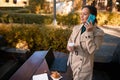 Dark-haired pretty woman with ponytail, in beige trench and green sweater, talks on mobile phone looking to the side, holds paper Royalty Free Stock Photo