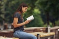 Young brunette sits on the bench and reads book. Trees at background Royalty Free Stock Photo