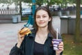 Young brunette with a smile holds a cheeseburger and a glass with a drink in her hands