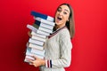 Young brunette student girl holding a pile of books afraid and shocked with surprise and amazed expression, fear and excited face Royalty Free Stock Photo