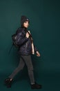 Young brunette teen girl, in jacket and hat with a bag, walking over dark-green background, Royalty Free Stock Photo