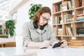 Young brunette teenager girl college student in glasses doing homework with mobile phone at modern library public place, green Royalty Free Stock Photo