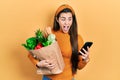 Young brunette teenager holding bag of groceries using smartphone celebrating crazy and amazed for success with open eyes Royalty Free Stock Photo