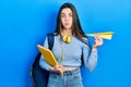 Young brunette teenager student holding books and paper plane puffing cheeks with funny face