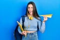 Young brunette teenager student holding books and paper plane winking looking at the camera with sexy expression, cheerful and Royalty Free Stock Photo