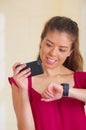 Young brunette wearing red top facing camera, holding mobile phone and looking at smart watch, white studio background Royalty Free Stock Photo