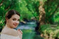 Young brunette woman alone with elegant hair bun in white off shoulder blouse enjoys nature green park with river smiling and look Royalty Free Stock Photo