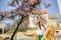 Young brunette woman artist holding in hands a brush and palette. Near her the magnolia tree and various art equipment