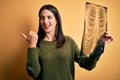 Young brunette woman with blue eyes holding xray of lungs over yellow background pointing and showing with thumb up to the side