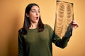 Young brunette woman with blue eyes holding xray of lungs over yellow background scared in shock with a surprise face, afraid and