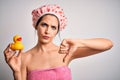 Young brunette woman with blue eyes wearing bath towel and shower cap holding duck toy with angry face, negative sign showing