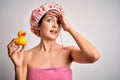 Young brunette woman with blue eyes wearing bath towel and shower cap holding duck toy stressed with hand on head, shocked with Royalty Free Stock Photo