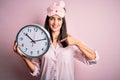Young brunette woman with blue eyes wearing pajama holding big clock at night time with surprise face pointing finger to himself Royalty Free Stock Photo